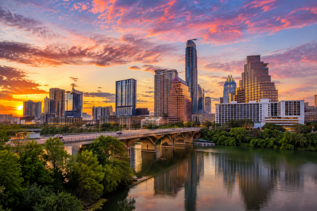 Austin, Texas skyline at sunset, with downtown skyscrapers, a bridge over the Colorado River, and colorful pink and orange clouds reflected in the water.
